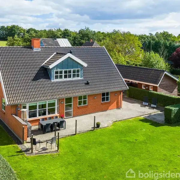 En rød murstensvilla med mørkt tag står på en velplejet grøn græsplæne omkranset af hække. Terrassen har havemøbler, og en garagebygning er synlig i baggrunden.