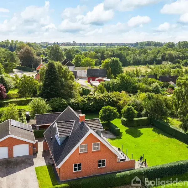 Et orange murstenshus med skråt tag står på en velplejet grøn græsplæne ved siden af en garage. Omgivelserne er frodige med træer og buske i et landskab med spredte huse.