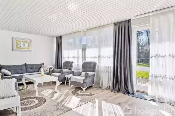 Stue med gråt møbelsæt og hvide og mørke gardiner, indrammet af lyse vægge. Stuen har et sofabord, sofagruppe og udsigt til terrasse gennem store vinduer.