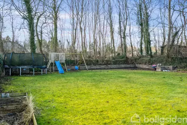 Trampolin og gyngestativ med rutsjebane står i en græsplæneomgivet have med træer i baggrunden.