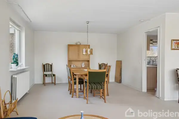 Et spisebord med seks stole står centralt i et lyst rum. En enkelt lampe hænger over bordet, og i baggrunden står et vitrineskab. Til højre ses en åben dør til køkkenet.