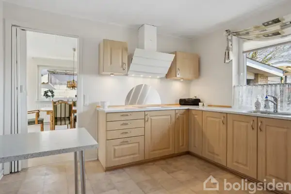 Et lyst køkken med træskabe har en kogeplade og emhætte. En bordplade strækker sig langs væggen, og et vindue giver udsigt til en baghave. I baggrunden ses en spisestue.