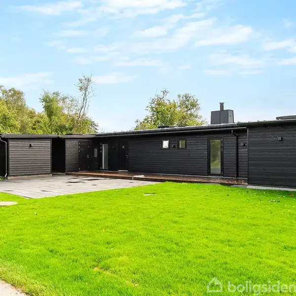 Et moderne sort træhus står stille på en velplejet grøn græsplæne, omgivet af høje træer under en skyfri himmel.