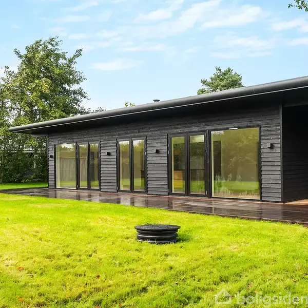 Et sortmalet, modernistisk hus med store vinduer står på en velholdt græsplæne omgivet af træer. Hustaget er fladt, og der er en terrasse langs facaden.