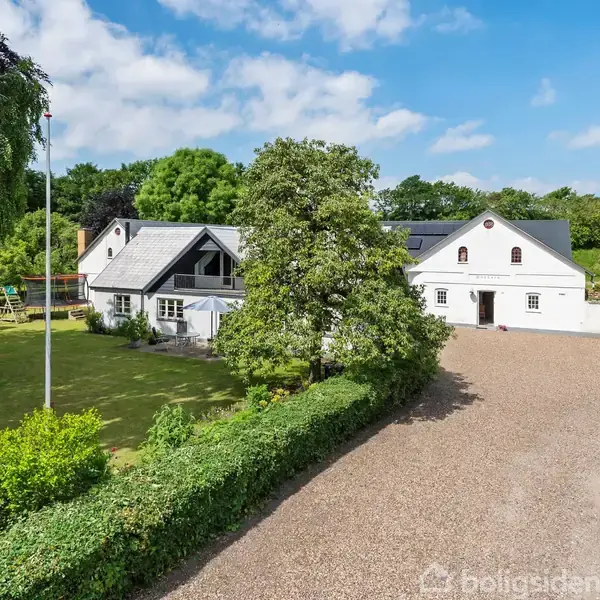 Et hvidt hus med gråt tag står ved siden af en grusvej omgivet af grønne træer og buske. En stor flagstang er placeret i haven.