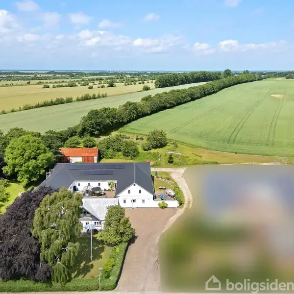 Gård med hvidt hus og sort tag omgivet af frodige marker og træer. Vejen strækker sig fra gården, mens det åbne landskab udstråler fred og landlig idyl.