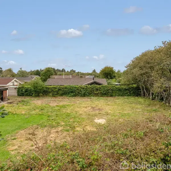 Græsplæne ligger ubenyttet med spredte buske og træer i forgrunden. I baggrunden ses et parcelhuskvarter med lave huse omgivet af hække. Skyet himmel i baggrunden.