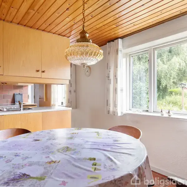 Et rundt bord med en blomstret dug står i et køkken-alrum, omgivet af træloft og et stort vindue med udsigt til en have. En lampe hænger over bordet.