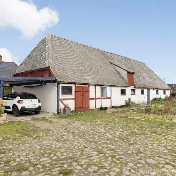 En stor gårdbygning med skråt tag står stille i et åbent gårdmiljø. En hvid bil holder parkeret under en blå træcarport ved siden af en del af bygningen.