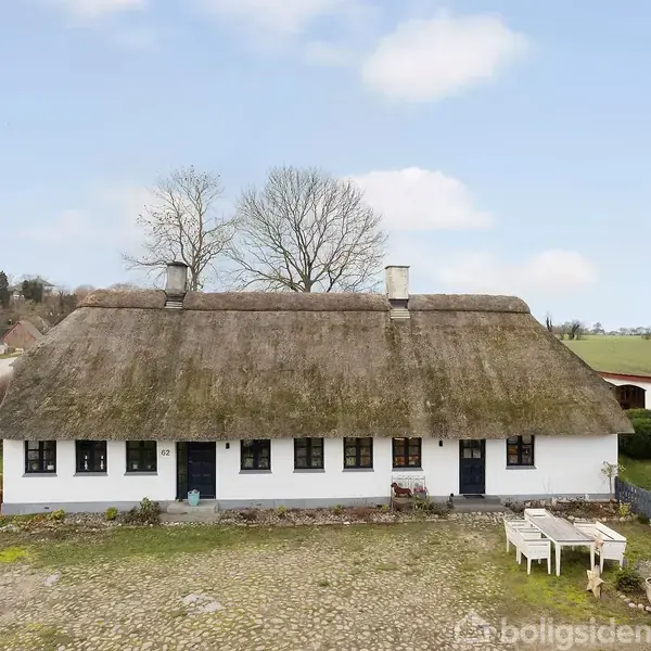 Stråtækt bindingsværkshus står stille på en brostensbelagt gårdsplads. Omkringliggende landskab består af åbne marker og spredte huse under en blå himmel med få skyer.