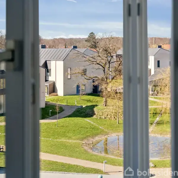 En udsigt gennem et vinduesparti viser en række moderne huse i et grønt boligområde med græsarealer, små stier og en lille dam.
