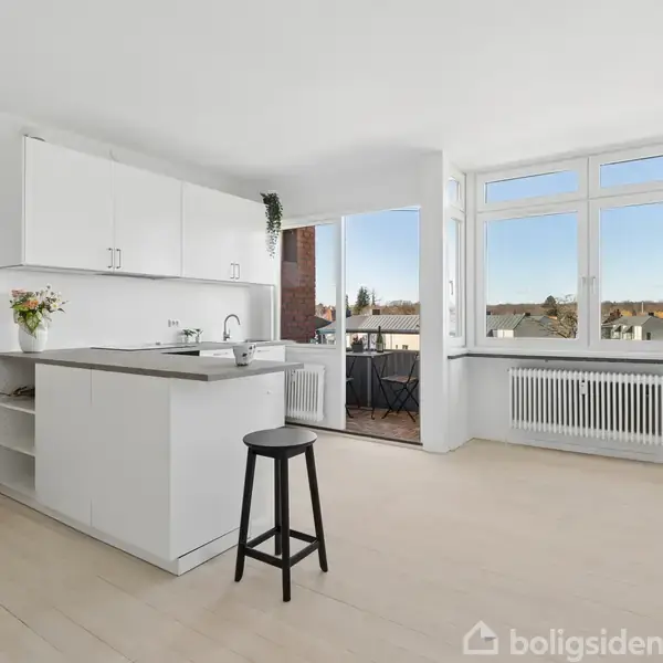 Køkkenet har hvide skabe og en høj barstol ved køkkenøen. Store vinduer med udsigt til en balkon og baggrund af huse og himmel. Blomster på bordet.