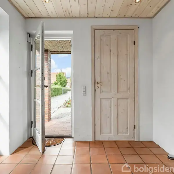 En lys entré med åben dør til en terrasse udenfor, omgivet af hvide vægge og et træloft med spotlys samt gulvfliser i terrakotta.