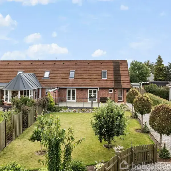 Et rødt murstenshus med mange vinduer og en sort tagkonstruktion står omgivet af en grøn have med stier og træer, omgivet af et hegn under en blå himmel med skyer.