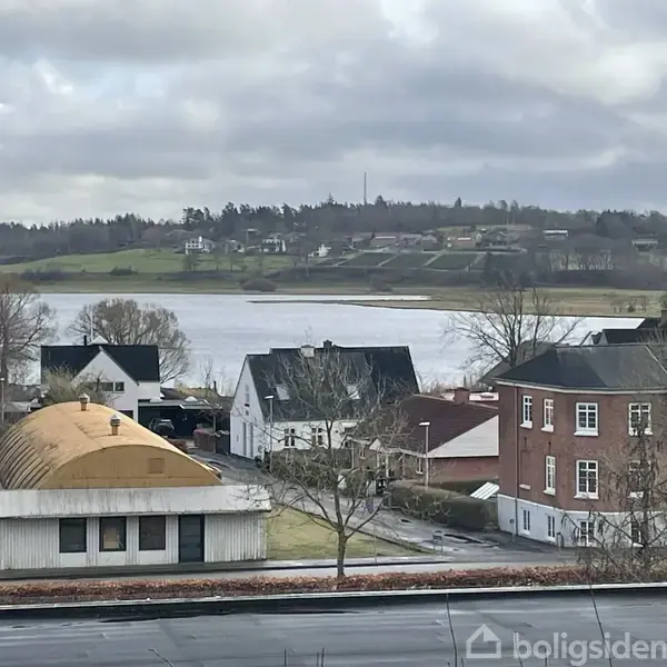 Et udsigt over et boligområde med huse i forgrunden, en flod i midten, og bakker med flere huse i baggrunden under en overskyet himmel.