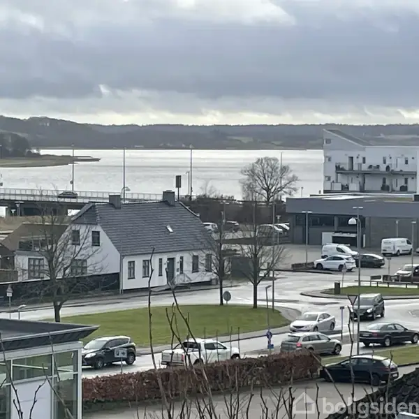 En hvid bygning med sadeltag står ved en vej. Biler kører forbi, mens en bro og vand ses i baggrunden under en overskyet himmel.