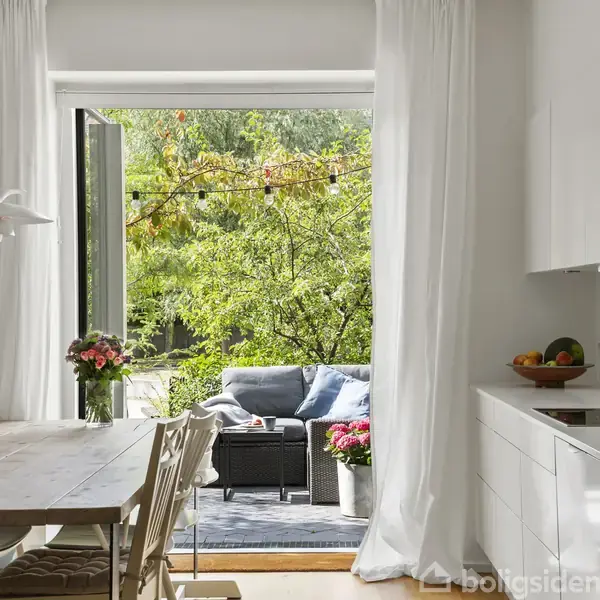 Et lyst køkken med et træbord og stole ved siden af skydedøre, der åbner op til en terrasse med sofa og blomster. Udsigt til grøn have med tæt løv.