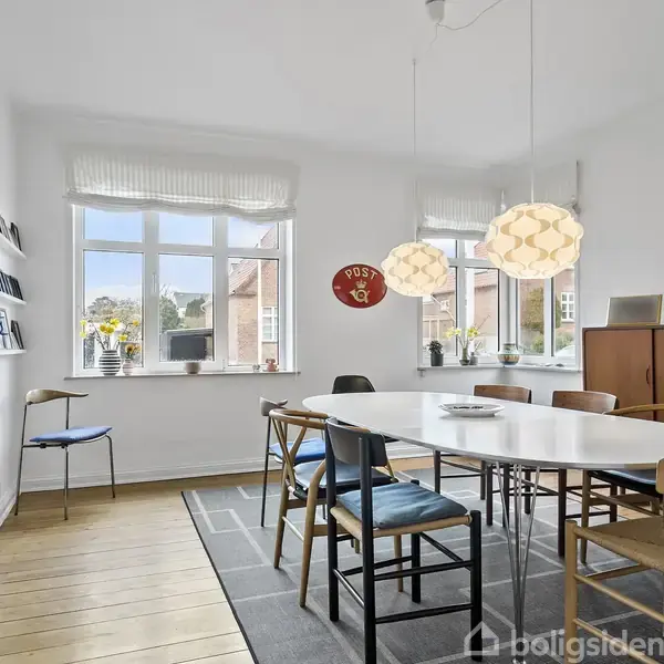 Et ovalt spisebord med seks stole står i et lyst rum med trægulv. Lamper hænger over bordet. Vinduer med udsigt til huse udenfor. En kommode står i baggrunden.