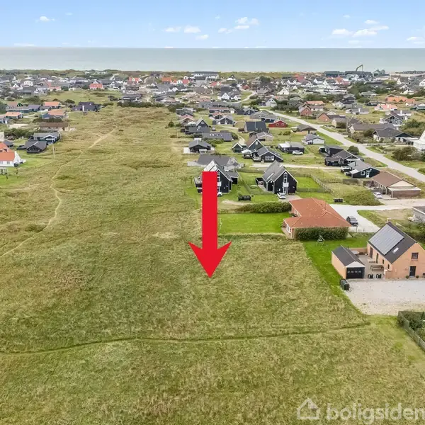 Rød pil peger mod en græsmark omgivet af huse i en åben landlig bebyggelse nær kysten med havet i baggrunden.