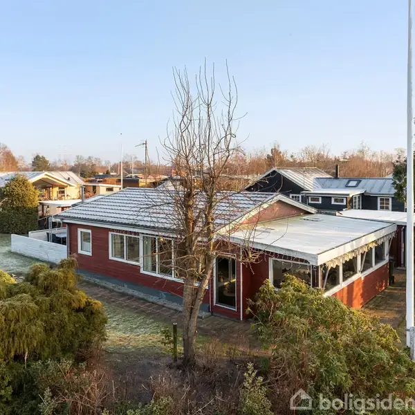 En rød murstensvilla med valmet tag står omgivet af grønne buske og et træ uden blade. Villaen har store vinduer og en overdækket terrasse i et villakvarter.