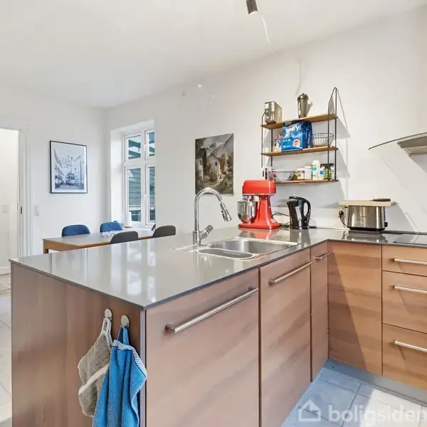 Køkken med træskabe, hvor en rød røremaskine står på bordet. I baggrunden er der en spiseplads og åben dør til en anden stue. Hvide vægge og hylder med genstande.