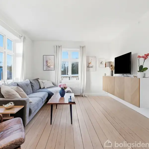 Stuen indeholder en grå sofa ved vinduer, et træbord med vase og fjernsyn på væghængt møbel. Lyse vægge og gulve skaber en lys og moderne atmosfære.