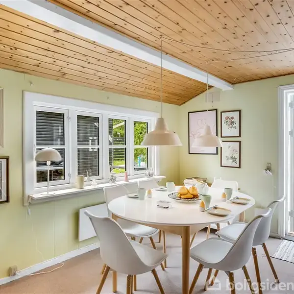 Et rundt spisebord med stole står dækket til morgenmad i et lyst spisekøkken med træloft. Store vinduer og en glasdør åbner ud til en terrasse med udsigt over en have.