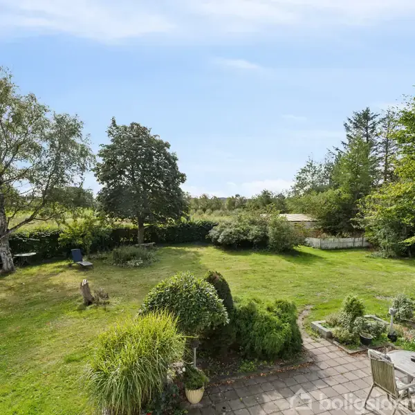 En frodig have med grønne plæner og træer omgiver en terrasse med havemøbler. Et hegn af planter markerer grænsen til resten af landskabet, som strækker sig mod horisonten.