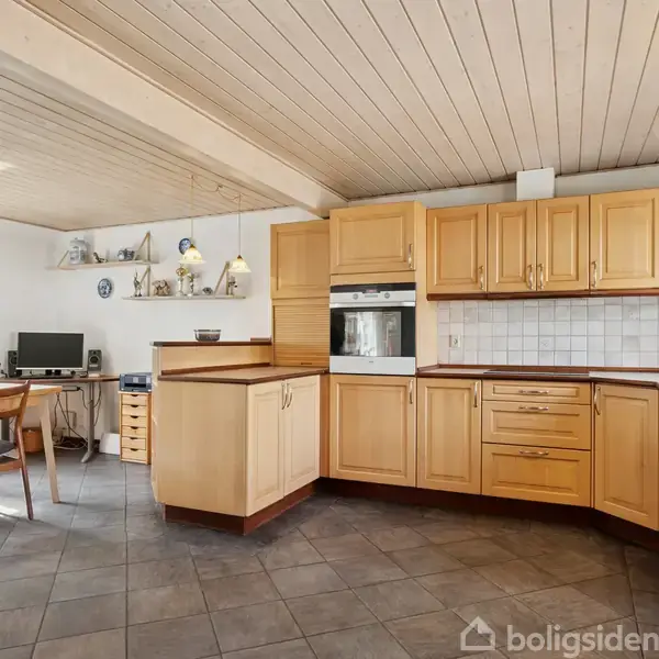 Et køkken med træskabe er omkranset af hvide vægge og et træloft. Der er et hjørne med en indbygget ovn, og et spiseområde med et bord og stole.