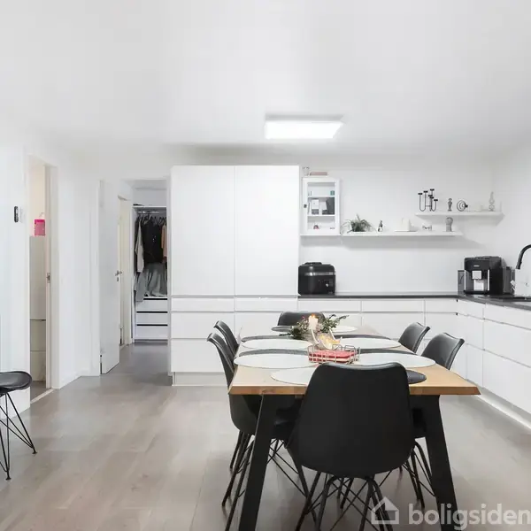 Et moderne køkken med et spisebord i centrum. Seks sorte stole omkranser bordet. Køkkenets hvide skabe og apparater er stilfuldt arrangeret, mens grønne planter tilføjer farve.