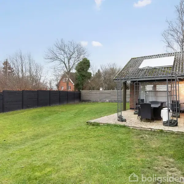 Terrasse med bord og stole under halvtag ved siden af et murstenshus. Græsplænen omgiver terrassen, og i baggrunden ses et sort plankeværk og bladløse træer.