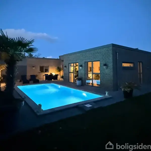 En moderne, firkantet bygning med store vinduer omgiver en oplyst swimmingpool om aftenen. Planter og havemøbler skaber en hyggelig atmosfære i baghaven.