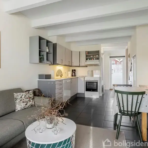 En grå sofa med et mønstret pude står i en stue ved siden af et hvidt sofabord med dekoration. Foran ses et lyst køkken med grå skabe og en spiseplads.