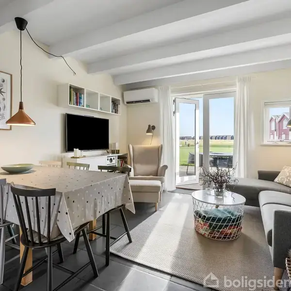 En hyggelig stue med spisebord dækket af en dug, en grå sofa ved siden af et sofabord, og store vinduer der åbner ud til en græsplæne udenfor.