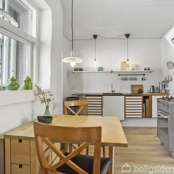 Trækøkkenbord med to stole står ved siden af et stort vindue. Bagved er der en køkkensektion med træskabe, hylder og diverse køkkenudstyr i rustfrit stål.