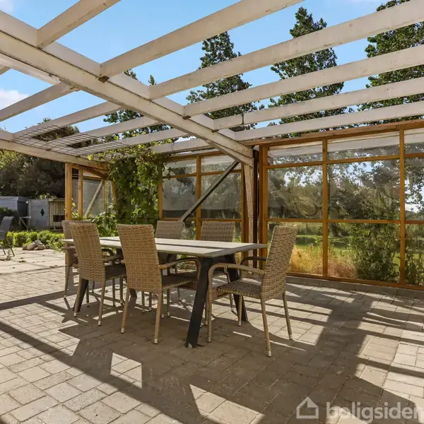 Et udendørs spisebord omgivet af seks stole står under en pergola med træramme. Omkring pergolaen er der netvægge, og landskabet i baggrunden består af en grøn mark og træer.