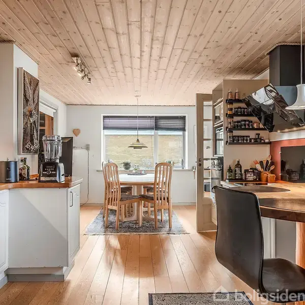 Køkken med hvide skabe, træbordplade og moderne apparater. Rundt spisebord med træstole i baggrunden. Trægulv og -loft skaber en hyggelig atmosfære. Ingen synlig tekst.