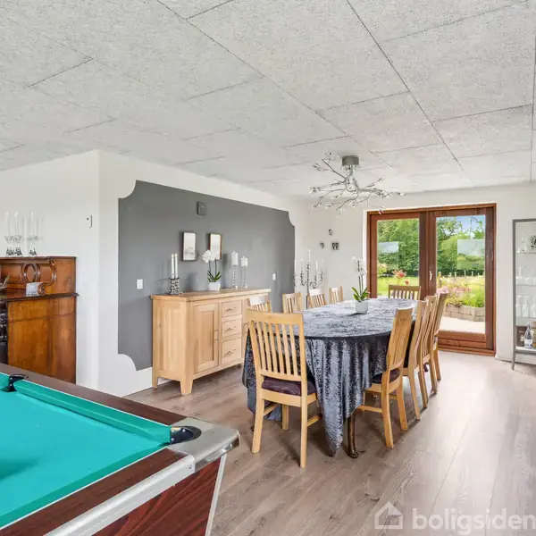 Poolbord står i stuen, klar til brug, med kugler trianguleret. Spisebord med dug og stole bagved. Store vinduer åbner ud til en grøn have.