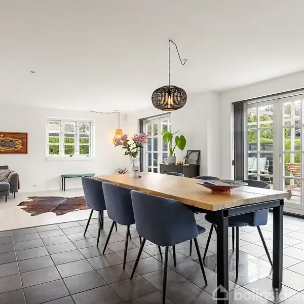 Et spisebord med blå stole står i et moderne, lyst rum med flisegulv. En lampe hænger over bordet, og der er en sofa og vinduer i baggrunden.