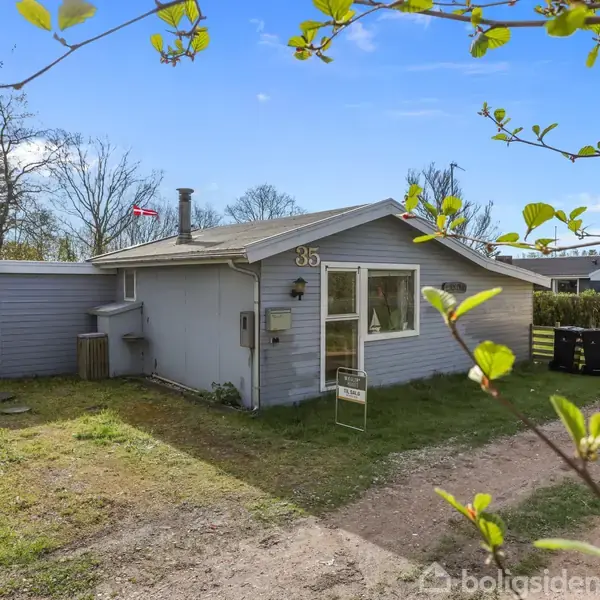 Gråmalet hus med nummer 35 ses fra vejen, omgivet af grønne buske og træer, under en blå himmel med et fåtal af skyer.