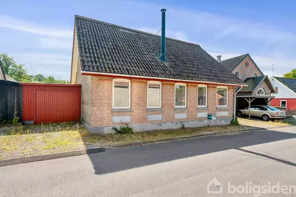 Et enfamiliehus står stille langs en rolig vej med en murstensfacade, skråt tag og en rød træport ved siden af. Solskin belyser det grønne område i baggrunden.