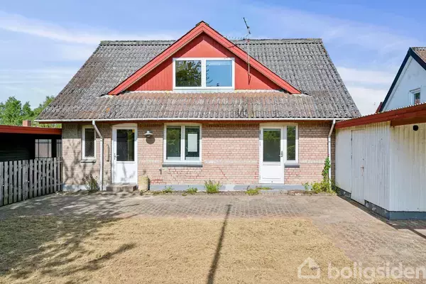 Et rødstenshus med to døre og flere vinduer står i en forhave med plæne og fliser. Der er en carport til højre, og himlen er blå.