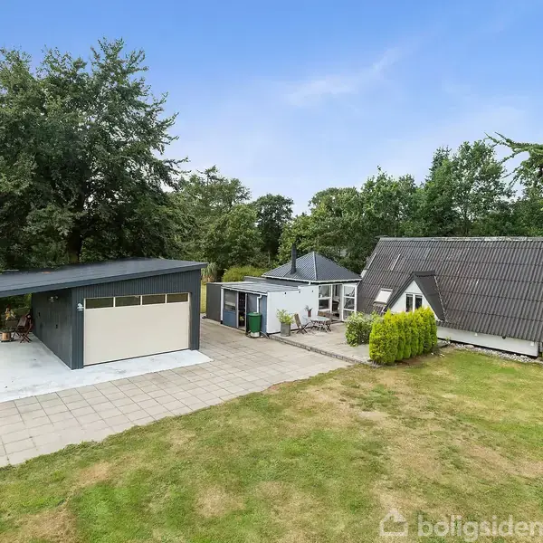 Et A-formet hus står ved siden af en moderne garage i en have omkranset af træer. En flisebelagt sti forbinder bygningerne, og omgivelserne er frodige med grønne græsplæner.