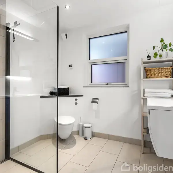 Badeværelse med bruseniche til venstre, toilet og papirkurv i midten. Hylde med planter, kurv og håndklæder til højre. Lys og moderne indretning med flisebelagt gulv.