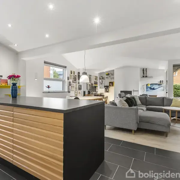 Køkkenø med moderne design står centralt i et åbent opholdsrum. Øen er omgivet af sofaarrangement, spisebord og store vinduer, der giver naturligt lys.