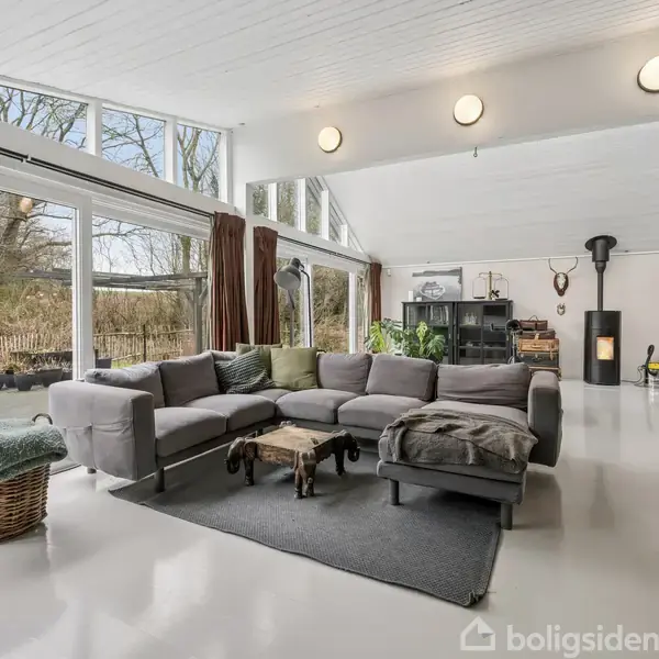 Gråt hjørnesofaarrangement vender mod vinduer i et lyst opholdsrum med moderne indretning. Pejs i hjørnet, tæppe under bord, og kurv med tæppe ved glasskydedøre ud mod have.