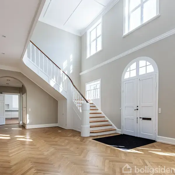 En elegant trappe fører op til første sal i et lyst og rummeligt indgangsparti med parketgulve og store vinduer, der giver rummet et indbydende og åbent udtryk.