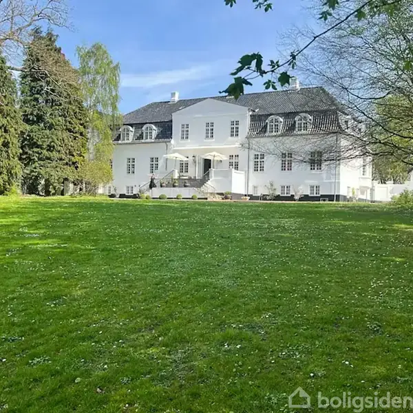Luftfoto af stor gård omgivet af træer og marker, markeret med orange linje. Baggrunden viser en sø og skovklædte områder under en klar blå himmel.