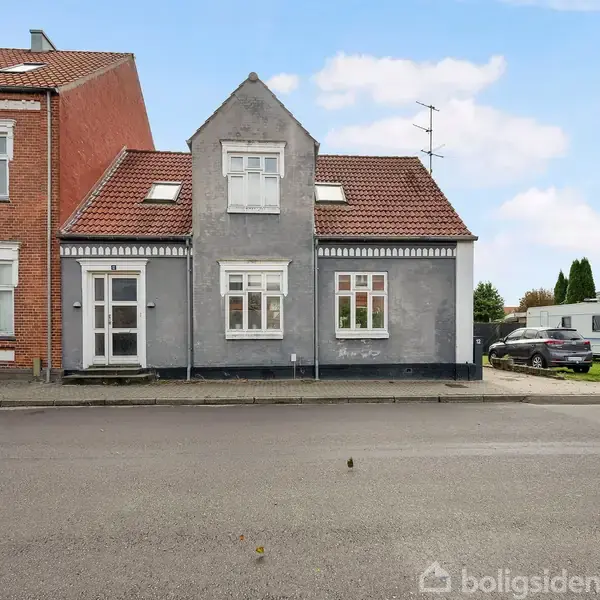 Et gråt hus med rødt tegltag står ved vejen. Ved siden af er en murstensbygning. En parkeret campingvogn og bil ses til højre, i en villavejsomgivelse.