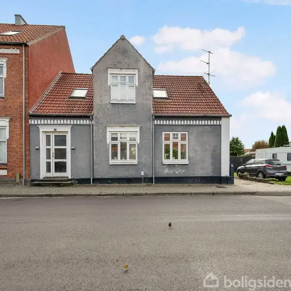 Et gråt hus med rødt tag står stille ved siden af et større rødt murstenshus. Foran er en vej, og til højre er en hvid campingvogn og bil parkeret.